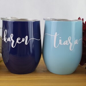 Puede incluir: Dos vasos de vino de acero inoxidable con tapas. El vaso de la izquierda es azul marino con el nombre "karen" escrito en blanco. El vaso de la derecha es azul claro con el nombre "tiara" escrito en blanco.