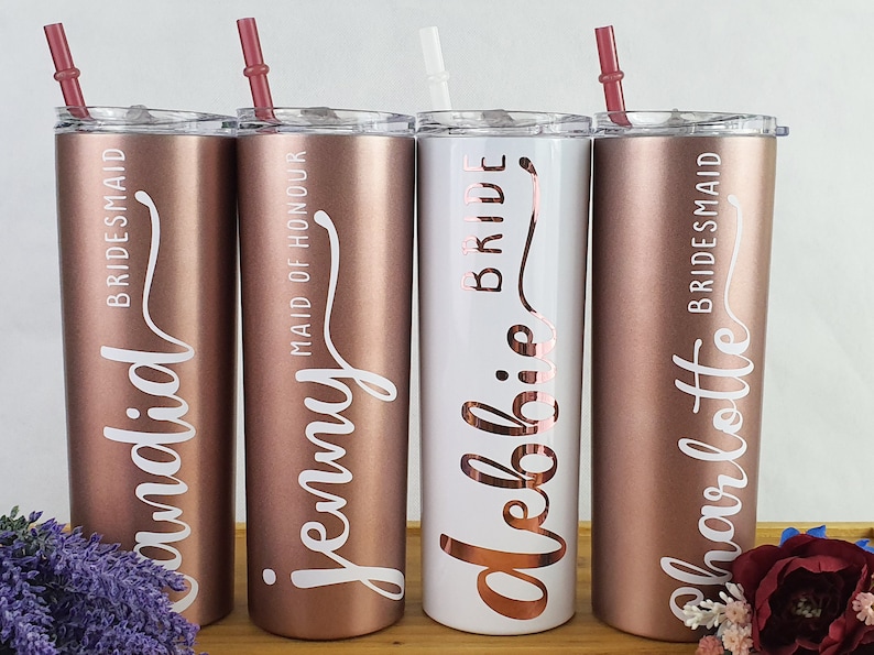 K&ouml;nnte beinhalten: Vier Edelstahlbecher mit ros&eacute;goldener Oberfl&auml;che und wei&szlig;er Beschriftung. Die Becher sind mit "Bride", "Maid of Honour" und "Bridesmaid" beschriftet, mit den Namen "Delohie", "Jenny" und "Charlotte".