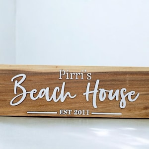 May include: A rectangular wooden sign with the words "Pirri's Beach House EST 2011" in white script. The sign is made of light brown wood. The text is centrally positioned on the sign.