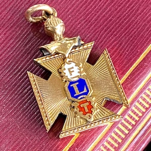 May include: A gold-toned pendant shaped like a cross with a textured sunburst pattern. The center features three stacked, decorative links with enamel accents: a white link with the letter F, a blue link with the letter L, and a red link with the letter T.