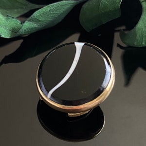 May include: A round, black and white brooch with a gold-colored metal rim. The brooch features a black background with a curved white stripe. The brooch is set against a dark, reflective surface.