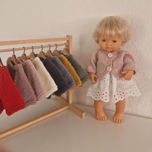Può includere: Una bambola con un cardigan rosa lavorato a maglia e un vestito bianco con occhielli è in piedi accanto a un attaccapanni di legno. L'attaccapanni espone una varietà di maglioni lavorati a maglia in miniatura in rosso, rosa, bianco, blu, giallo e verde.