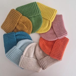 Könnte beinhalten: Ein Set aus zehn gestrickten Mützen in verschiedenen Farben, darunter Senfgelb, Blaugrün, Hellblau, Creme, Orange, Rosa und Grün. Die Mützen sind in einem Kreis angeordnet, wobei die Oberseiten der Mützen nach außen zeigen.
