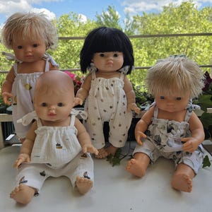 Peut inclure: Quatre poupées pour bébés avec différentes coiffures et tenues. Les poupées ont la peau claire et portent des barboteuses blanches avec divers motifs, notamment des citrons, des fleurs et des bateaux. Elles sont posées sur une surface blanche avec un fond vert.