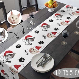 May include: White table runner with a Christmas cat theme. The runner features a repeating pattern of paw prints, cats wearing Santa hats, and the names "Luna" and "Foxy". The text "Up to 5 Cats" is printed at the bottom of the runner.