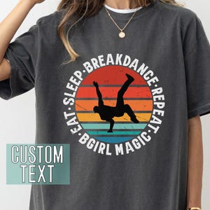Puede incluir: Una camiseta gris oscuro con un gráfico de estilo vintage de un breakdancer en un círculo. El texto alrededor del círculo dice "Eat. Sleep. Breakdance. Repeat. Bgirl Magic."