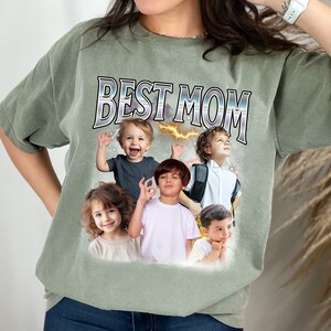 K&ouml;nnte beinhalten: Ein gr&uuml;nes T-Shirt mit dem Text "BEST MOM" in silbernen Buchstaben. Das Shirt zeigt eine Collage aus f&uuml;nf Kindergesichtern.