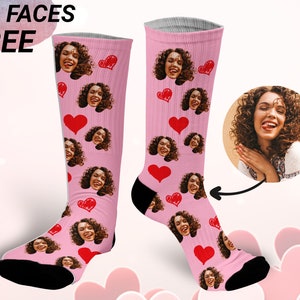 May include: A pair of pink socks with a heart pattern and multiple faces printed on them. The socks are knee-high and have a black toe and heel. The text "1-5 FACES FREE" is visible in the top left corner of the image.