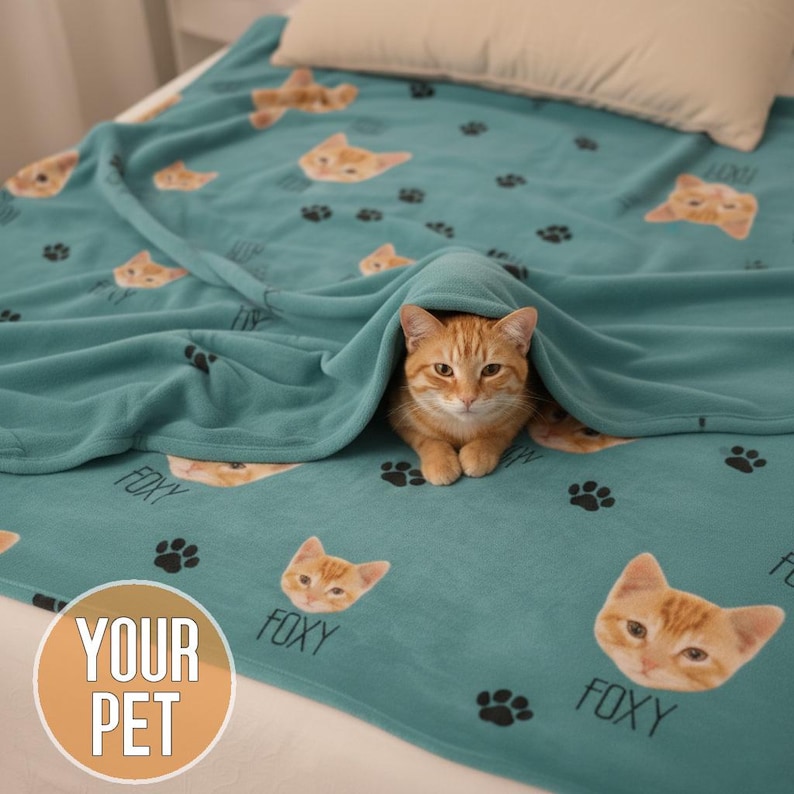 A person holds up a teal blanket featuring multiple ginger cat faces, each labeled with black paw prints scattered between them. A real orange tabby cat sits in front of the blanket, and a small snow-dusted Christmas tree stands nearby on a bright wooden floor against a white wall.