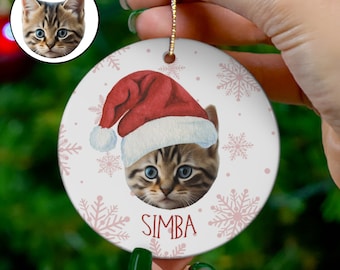 May include: A white ceramic Christmas ornament with a cute cat wearing a red Santa hat. The cat's name, "SIMBA", is printed in pink below the cat's face. The ornament is decorated with pink snowflakes and white dots.