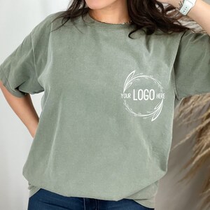 May include: A sage green t-shirt with a white floral design and the text "YOUR LOGO HERE" in the center.