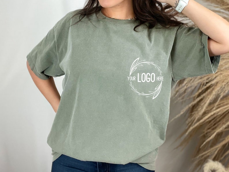 May include: A sage green t-shirt with a white floral design and the text "YOUR LOGO HERE" in the center.