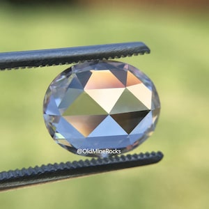 May include: A clear, faceted, oval-shaped gemstone with a rose cut. The gemstone is held in tweezers against a green background.