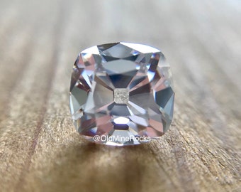 May include: A clear, square-cut gemstone with a central square facet. The gem has multiple facets that reflect light, creating a sparkling effect. The background is a wooden surface. The text "@OldMineRocks" is visible in the lower left corner.