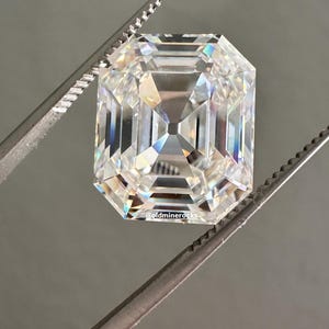 May include: A large, clear, emerald-cut gemstone held by a pair of tweezers. The gemstone has multiple facets that reflect light, creating a sparkling effect. The background is a solid, neutral gray.