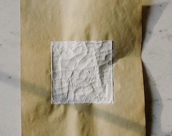Puede incluir: Un cuadrado de material blanco texturizado está adherido a una hoja de papel marrón. El material blanco tiene una superficie agrietada e irregular. El papel es de color marrón claro. El fondo es una superficie de mármol blanco.