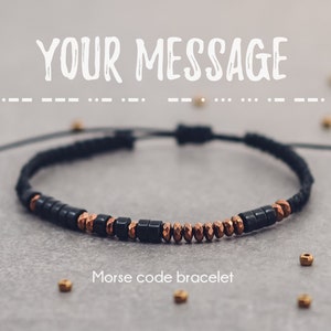 Può includere: Un braccialetto con perline nere e rame con un cordoncino nero. Il braccialetto ha il testo "YOUR MESSAGE" sopra e "Morse code bracelet" sotto.