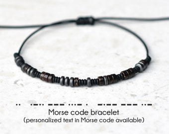 Può includere: Un braccialetto in corda nera con perline marroni e argentate. Le perline sono disposte in uno schema che rappresenta il codice Morse. Il testo sotto il braccialetto recita "Braccialetto in codice Morse (testo personalizzato in codice Morse disponibile)".