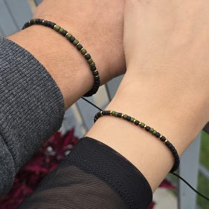 Peut inclure: Deux bracelets de perles sont présentés, un sur chaque poignet. Les bracelets comportent des perles noires, vert olive et dorées enfilées sur un cordon noir. Les bracelets sont portés par deux personnes, leurs poignets se touchant.