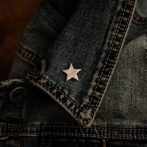 May include: A small, five-pointed silver star pin attached to the collar of a blue denim jacket. The jacket features visible dark blue stitching along the seams and a metallic button near the collar, showcasing a textured fabric appearance.