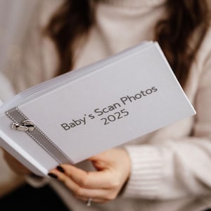 Könnte beinhalten: Ein weißes Fotoalbum mit einem silbernen Band und einem silbernen Anhänger mit zwei Fußabdrücken. Der Albumcover hat den Text "Baby's Scan Photos 2025" in grau gedruckt.