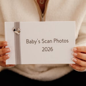 Pode incluir: Uma caixa branca com o texto "Baby's Scan Photos 2026" em cinza. Uma fita cinza com um berloque prateado em forma de pegadas de bebê decora a caixa. A caixa é segurada por uma pessoa.