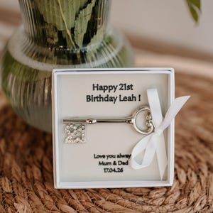 Puede incluir: Una llave plateada con un dije '21' en una caja blanca. La caja tiene el texto 'Happy 21st Birthday Leah!' y 'Love you always Mum & Dad 17.04.26'. Una cinta blanca está atada a la caja. El fondo es una estera tejida.