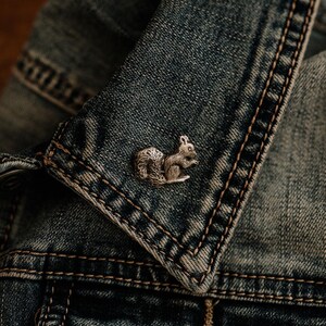 May include: A silver-toned metal pin shaped like a squirrel with detailed fur texture, attached to the collar of a blue denim jacket. The squirrel is depicted in a seated position with its bushy tail curved upward, showcasing a metallic finish.