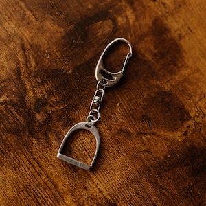 Puede incluir: Llavero plateado con diseño de estribo. El llavero tiene un cierre para una fácil fijación y una pequeña cadena que conecta el estribo al cierre. El fondo es una superficie de madera marrón oscuro.