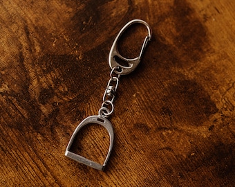 May include: Silver-coloured keychain featuring a stirrup design. The keychain has a clasp for easy attachment and a small chain connecting the stirrup to the clasp. The background is a dark brown wooden surface.