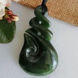 May include: A dark green jade pendant with a spiral design, suspended from a black cord. The pendant is smooth and polished. The image includes white flowers with red accents and a small burlap bag.