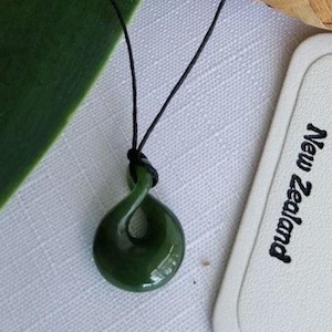 Op de afbeelding: Een donkergroene, gepolijste jade hangerketting met een gedraaid, spiraalvormig ontwerp. De hanger hangt aan een zwart koord. Een wit label met "New Zealand" is zichtbaar op de achtergrond.