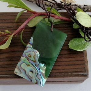 May include: A rectangular green pendant with an abalone shell inlay, suspended from a brown cord. The pendant is displayed on a wooden box, with green leaves and branches in the background. The abalone shell has iridescent colors.