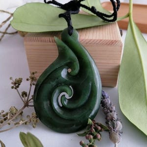May include: A dark green jade pendant with a spiral design, suspended from a black cord. The pendant is set against a wooden box and surrounded by greenery and dried flowers, creating a natural aesthetic.