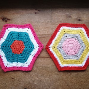 May include: Two crocheted hexagonal placemats with colorful geometric patterns. One placemat features a turquoise center with a red circle, surrounded by white and pink borders. The other placemat has a pink center, surrounded by gray, yellow, white, and red borders.