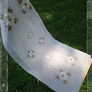 May include: A white linen table runner with embroidered brown and white flowers. The runner is folded and draped over a wooden chair.