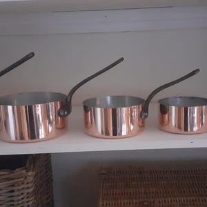 Peut inclure: Ensemble de cinq casseroles en cuivre avec des poignées noires. Les casseroles sont disposées par ordre de taille, la plus grande à gauche et la plus petite à droite.