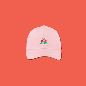May include: A light pink baseball cap with a curved brim. The cap features a red rose with green leaves embroidered on the front. The hat is set against a solid orange background.