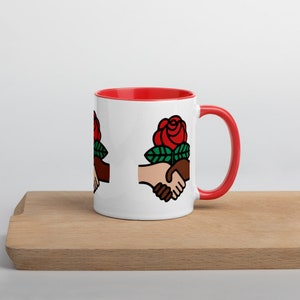 Puede incluir: Taza de cerámica blanca con interior y asa rojos. La taza presenta un gráfico de dos manos estrechándose, con una rosa roja y hojas verdes encima. La taza está sobre una superficie de madera.