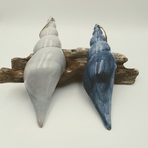 May include: Two ceramic seashell ornaments, one in a light gray color and the other in a dark blue color. The seashells are hanging from a piece of driftwood.