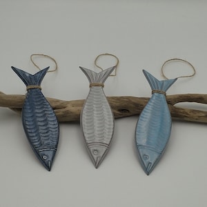 Puede incluir: Tres adornos de pescado de cerámica colgando de un cordel. Los peces son en tonos de azul, gris y blanco. Los peces están colgando de un trozo de madera a la deriva.