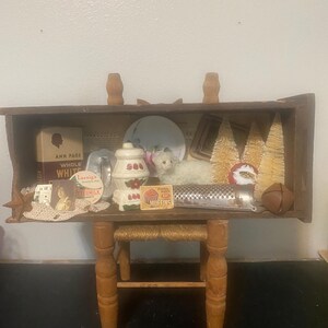 May include: A miniature wooden shelf with a white lamb, a small red and white ceramic teapot, a grater, and a box of muffins. The shelf is on a small wooden stool with a rope seat.