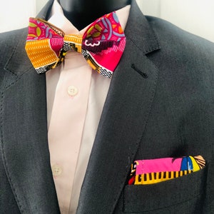 Può includere: Un set di papillon e pochette fantasia. Il papillon presenta un mix di colori tra cui rosa, giallo e blu. La pochette è nella tasca della giacca. L'abito è grigio e la camicia rosa chiaro.