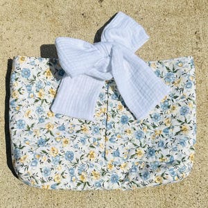 May include: A rectangular tote bag with a floral pattern in blue and yellow on a white background. A large white bow is tied on top of the bag. The bag is made of fabric.