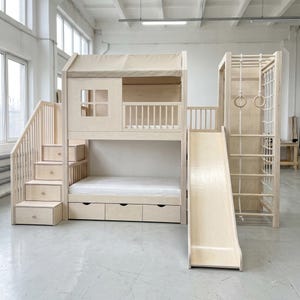 May include: A light-coloured wooden bunk bed structure featuring a house-shaped upper loft with a window and railing, a lower bed with three storage drawers, a staircase with built-in drawers, a slide, and a climbing frame with rings and a net.
