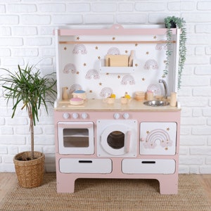 May include: Pink wooden play kitchen with a white countertop, oven, and sink. The backsplash features a rainbow and star pattern. The kitchen includes a variety of accessories, such as pots, pans, and utensils.