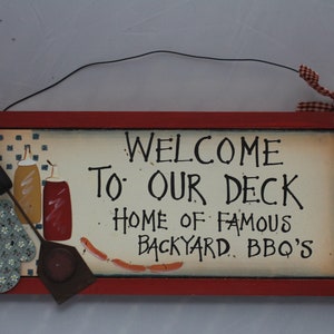 May include: A red wooden sign with white lettering that reads "Welcome to our deck, Home of famous backyard BBQ's". The sign has a black wire hanger and a red and white checkered ribbon. The sign is decorated with a spatula, a blue and white patterned oven mitt, and two bottles of condiments.