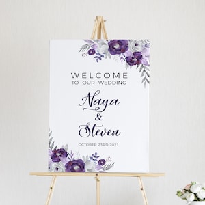 May include: A white wedding welcome sign with purple and gray watercolor flowers. The sign reads "Welcome to our wedding, Naya & Steven, October 23rd 2021".