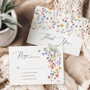 May include: Two white cards with floral designs. The top card says "Thank You" with a floral border. The bottom card says "RSVP by June 28th, 2021" with a floral design and a dragonfly. The cards are on a white lace fabric.