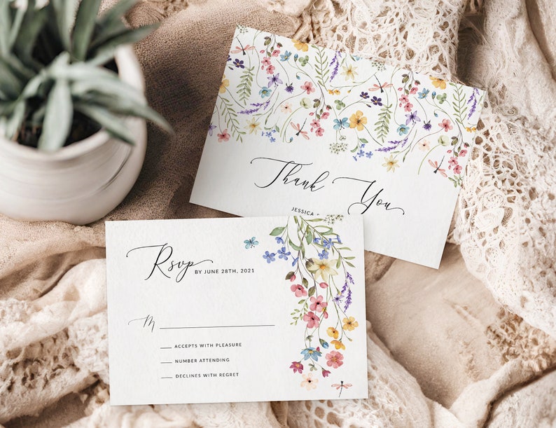 May include: Two white cards with floral designs. The top card says "Thank You" with a floral border. The bottom card says "RSVP by June 28th, 2021" with a floral design and a dragonfly. The cards are on a white lace fabric.